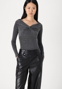 Maglietta a maniche lunghe grigio metallizzato con design anteriore attorcigliato, abbinata a pantaloni di pelle nera a vita alta con grandi bottoni decorativi.