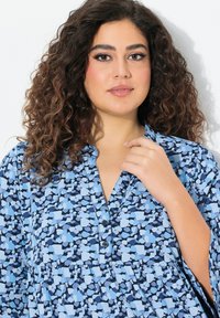 Blusa a fantasia blu con colletto a notched e bottoni. Il design presenta tonalità variabili di blu in forme astratte su uno sfondo chiaro.