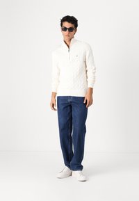Witte kabelgebreide trui met een ritskraag, gecombineerd met blauwe spijkerbroek en witte sneakers, model draagt donkere zonnebril.