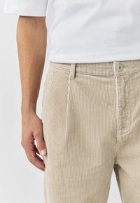 Beige corduroy trousers with vertical ribbed texture, a front button closure, and side pockets. Simple design with a tailored fit.