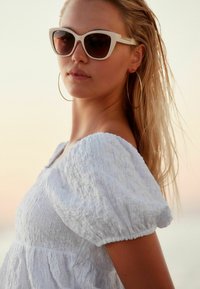 Młoda kobieta z mokrymi blond włosami nosi białą, fakturowaną bluzkę z bufkami, duże kolczyki w kształcie kół oraz białe okulary w stylu kocich oczu o zachodzie słońca.