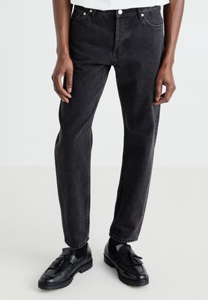 Person iført sorte jeans med lige ben, sorte loafers med kvaster og en hvid skjorte foran en ensfarvet lys baggrund.