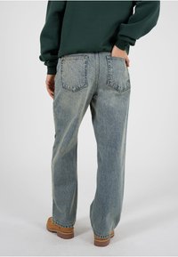 Persoană îmbrăcată în blugi albaștri deschiși, cu croială dreaptă, hanorac verde închis și bocanci maro deschis cu șireturi, stând cu o mână în buzunarul din spate.