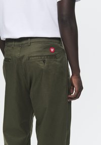 Olive green trousers with a tailored cut, featuring a rear pocket with a black button and a red circular logo patch.
