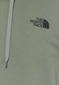Světle zelená mikina s předním zipem, texturovaná tkanina a černé logo "The North Face" na levé straně.