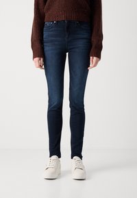 Skinny jeans in dark wash denim, featuring a mid-rise waist and a smooth texture. Paired with white sneakers and a brown knitted sweater.