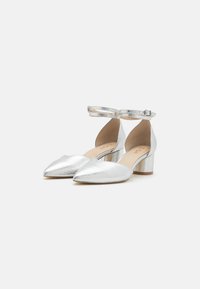 Chaussures à talons aiguilles métalliques argentées avec un bout pointu, une chaîne à la cheville, un design ouvert à l'arrière et un talon épais. Texture lisse et réfléchissante.