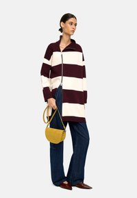Bordowy i kremowy sweter w paski o oversize'owym kroju z zapięciem na zamek z przodu, w połączeniu z ciemnoniebieskimi spodniami o szerokich nogawkach i małą żółtą torebką z zamszu.