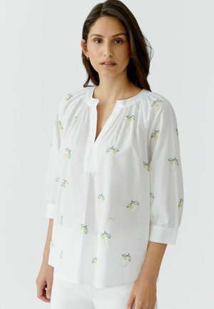 Frau mit langen dunklen Haaren, die eine weiße Bluse mit gelber Zitronenstickerei und Dreiviertelärmeln vor einem einfarbigen hellen Hintergrund trägt.