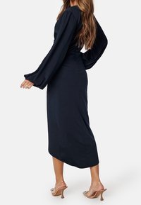 Long-sleeve, navy blue dress in smooth fabric with a fitted waist and flowing hem, paired with light brown heeled sandals.