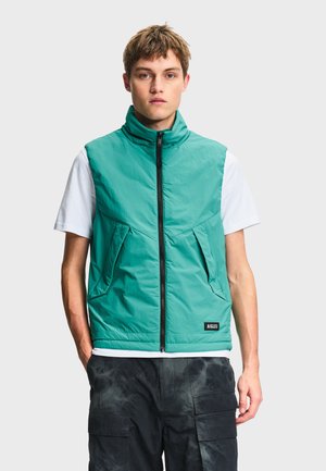 Jeune homme portant un gilet sans manches zippé couleur sarcelle par-dessus un t-shirt blanc et un pantalon cargo noir, faisant face à l'avant sur un fond uni.