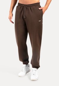 Braune Sweatpants aus weichem Material, mit einem elastischen Bund, Bündchen an den Knöcheln und Seitentaschen. Kombiniert mit weißen Turnschuhen.
