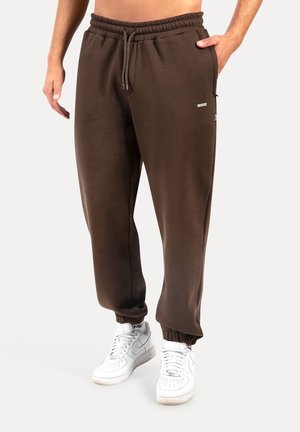 Braune Sweatpants aus weichem Material, mit einem elastischen Bund, Bündchen an den Knöcheln und Seitentaschen. Kombiniert mit weißen Turnschuhen.
