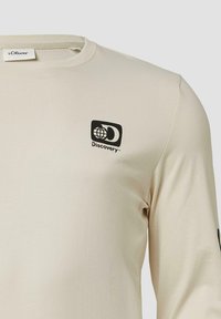 Beige langslijfshemd van gladde stof, met een zwart Discovery-logo op de linkerkant van de borst en een ronde halslijn.