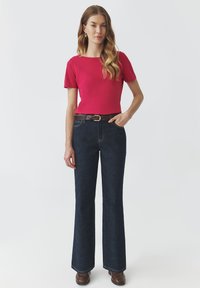Haut rouge à manches courtes associé à un jean évasé bleu foncé et une ceinture en cuir marron. La tenue est complétée par des bottines marron.