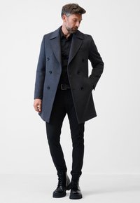 Grå uld dobbeltknapsjakke med store klapskuldre, sorte knapper og sidelommer. Stylet med sorte tætsiddende bukser og snørebåndsorter.