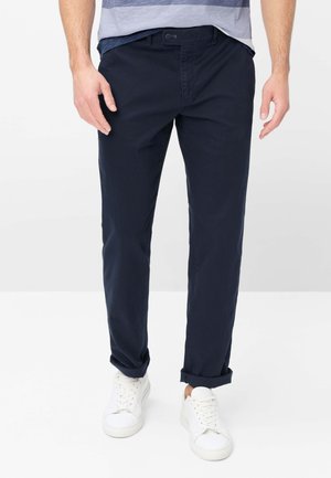 Mand iført mørkeblå op rullede bukser, hvide sneakers og en blå-grå stribet afslappet skjorte mod en hvid baggrund.