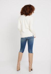 Valkoinen pörröinen neule pyöreällä kaula-aukolla, yhdistettynä sinisiin farkkuihin ja beigen värisiin polvimittaisiin saappaisiin, joissa on sileä pinta ja vähäistä kulumista.