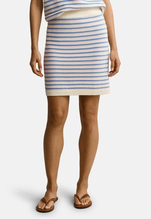 Frau trägt einen blau-weiß-pink gestreiften, gestrickten Minirock mit passendem Oberteil und braunen Flip-Flop-Sandalen vor einfachem Hintergrund.