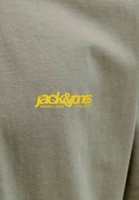 Tissu vert clair avec un petit logo texte jaune indiquant "jack & jones PROJECT CORE CREATIVE DEPT" sur la poitrine.