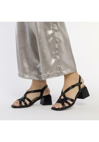 Sandales noires à bride avec des talons carrés, dotées de brides tressées et d'une boucle à la cheville. Portées avec un pantalon large argent métallique.