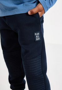 Marineblaue Jogginghosen mit strukturierten seitlichen Einsätzen und dem Aufdruck "PLAY ALL DAY". Hergestellt aus weichem Material, mit einer taillierten Passform und elastischem Bund.