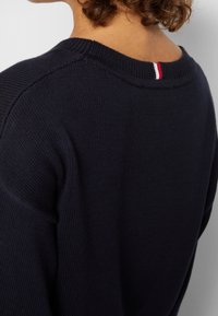 Personne aux cheveux bouclés portant un pull en maille bleu marine foncé avec un petit détail rayé rouge, blanc et bleu au dos de l'encolure.