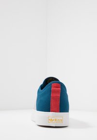 Modré plátěné tenisky s červeným patníkem a bílou podrážkou, pohled zboku zezadu, na bílé ploše s logem "Nizza" na podrážce.