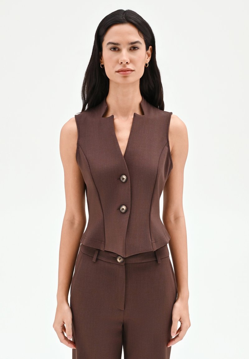 Donna con capelli lunghi e scuri che indossa un gilet marrone su misura senza maniche con due bottoni e pantaloni marroni coordinati.