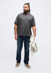 Άνδρας με dreadlocks που φοράει σκούρο γκρι μπλουζάκι, μπλε τζιν, καφέ αθλητικά παπούτσια, κρατώντας ένα διπλωμένο φωτεινό πουκάμισο με μοτίβο στο δεξί χέρι.