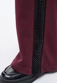 Donkerrode joggingsbroek met een zwart geweven accent langs de zijkant. De zoom heeft een contrasterende textuur. Zwarte glanzende schoenen zijn gedeeltelijk zichtbaar.