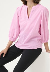 Blouse rose texturée avec un décolleté en V partiel et des manches ballon, associée à un pantalon noir. Le tissu présente un motif subtil.