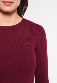 Pull en mailles côtelées bordeaux avec un col rond et des manches longues, mettant en valeur une texture douce et des lignes verticales à travers le tissu.