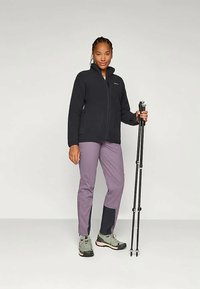 Μαύρο φλις μπουφάν με ψηλό κολάρο, σε συνδυασμό με μοβ παντελόνι πεζοπορίας που διαθέτει μαύρες λεπτομέρειες. Το μοντέλο κρατάει μπαστούνια πεζοπορίας και φοράει γκρίζα παπούτσια.