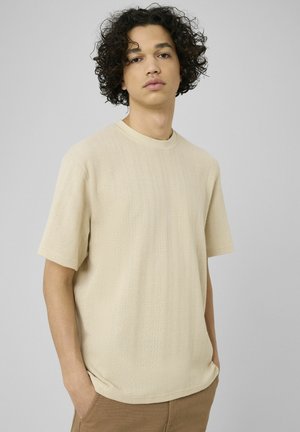 Jeune homme aux cheveux bouclés portant une chemise beige texturée à manches courtes et un pantalon marron, posant devant un fond gris uni.