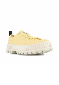 Zapatilla de lona amarilla con una suela gruesa y texturizada en blanco, presenta una lengüeta negra en el talón y cordones redondos. Diseño minimalista y material duradero.