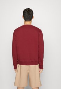 Felpa bordeaux con scollo a girder, polsini e orlo a costine. La vista posteriore mostra una texture liscia, abbinata a pantaloni corti beige.