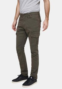 Pantalons cargo vert foncé avec de multiples poches, détails en zip et une coupe slim, associés à des baskets noires et un t-shirt gris.