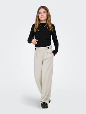 Fille aux longs cheveux portant un haut noir à manches longues, un pantalon beige à jambes larges avec des détails de boutons, des chaussures noires, et un collier coloré en perles, debout sur un fond blanc.