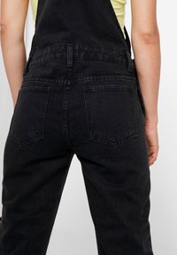 Svart denimoverall med en avslappnad passform, featuring en hög midja, två bakfickor och justerbara remmar. Slät, matt textur.