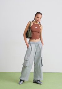 Brun, ermeløs crop top og grå vidt skjørt sweatpants med sidelommer. Bærer en grønn skulderveske, sølv sneakers på et grønt gulv.
