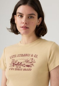 T-shirt beige a maniche corte con scollatura rotonda, caratterizzato da una grafica bordeaux di montagne e due cavalli, etichettato "LEVI STRAUSS & CO."