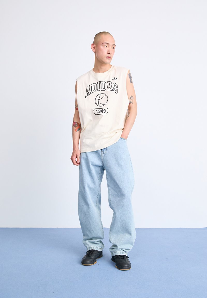 Camisa sin mangas de algodón beige con el logo de "ADIDAS" y un gráfico de baloncesto, combinada con jeans sueltos de color azul claro y zapatillas negras.