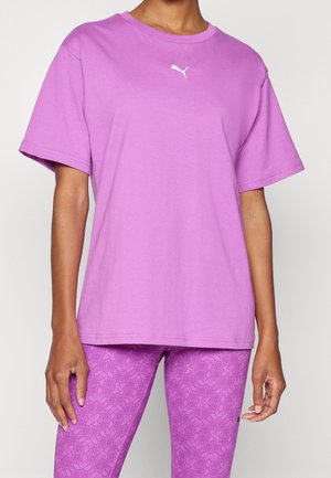 Osoba ubrana w jednolity liliowy T-shirt z krótkim rękawem z białym logo Puma oraz wzorzyste liliowe legginsy na neutralnym tle.