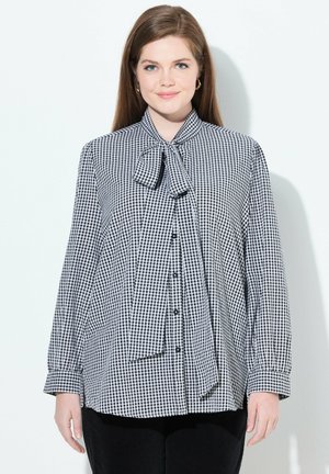 Blusa a quadri bianca e nera con colletto a fiocco, dotata di maniche lunghe e chiusura a bottoni. Il materiale sembra morbido e texturizzato.