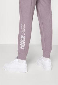 Pantaloni della tuta viola con testo bianco "NIKE AIR" sul lato, abbinati a scarpe da ginnastica bianche. I pantaloni presentano polsini a coste alle caviglie.