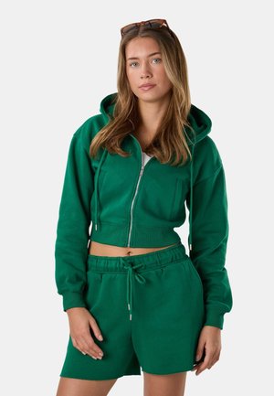 Young woman with long light brown hair wearing sunglasses on her head, a green cropped zip hoodie, and matching shorts.