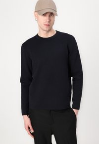 Marineblauer Strickpullover mit langen Ärmeln und Rundhalsausschnitt, kombiniert mit schwarzen Hosen, getragen unter einer beigen Baseballmütze.