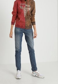 Bluza z kapturem w czerwono-brązowe bloki kolorystyczne z nadrukiem graficznym, zestawiona z niebieskimi dżinsami i białymi trampkami z wielokolorowymi akcentami.