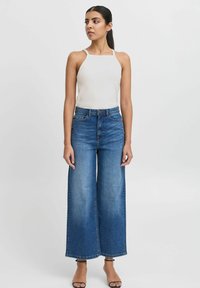 Top blanco con cuello halter acanalado combinado con jeans de mezclilla azul claro de pierna ancha. Los jeans tienen una cintura alta y un dobladillo limpio. Sandalias negras de tacón.